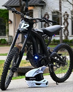 SURRON Light Bee X Electric Bike 2024, also known as Surron X, with a helmet placed in front of it. Key details include: It's an electric dirt bike, blending features of a mountain bike and a dirt bike.  The bike is lightweight at 104 lbs and powered by a 60V battery.  It has a top speed of 45 mph and a range of 40-60 miles.  The battery can last up to 80 miles on a single charge, depending on usage.  It features spoke wheels, front/rear 19 x 1.4 alloy hubs, and 70/100-19 CST tires.  The bike is suitable for both beginners and experienced riders.  It can be made street legal with the addition of mirrors and registration.